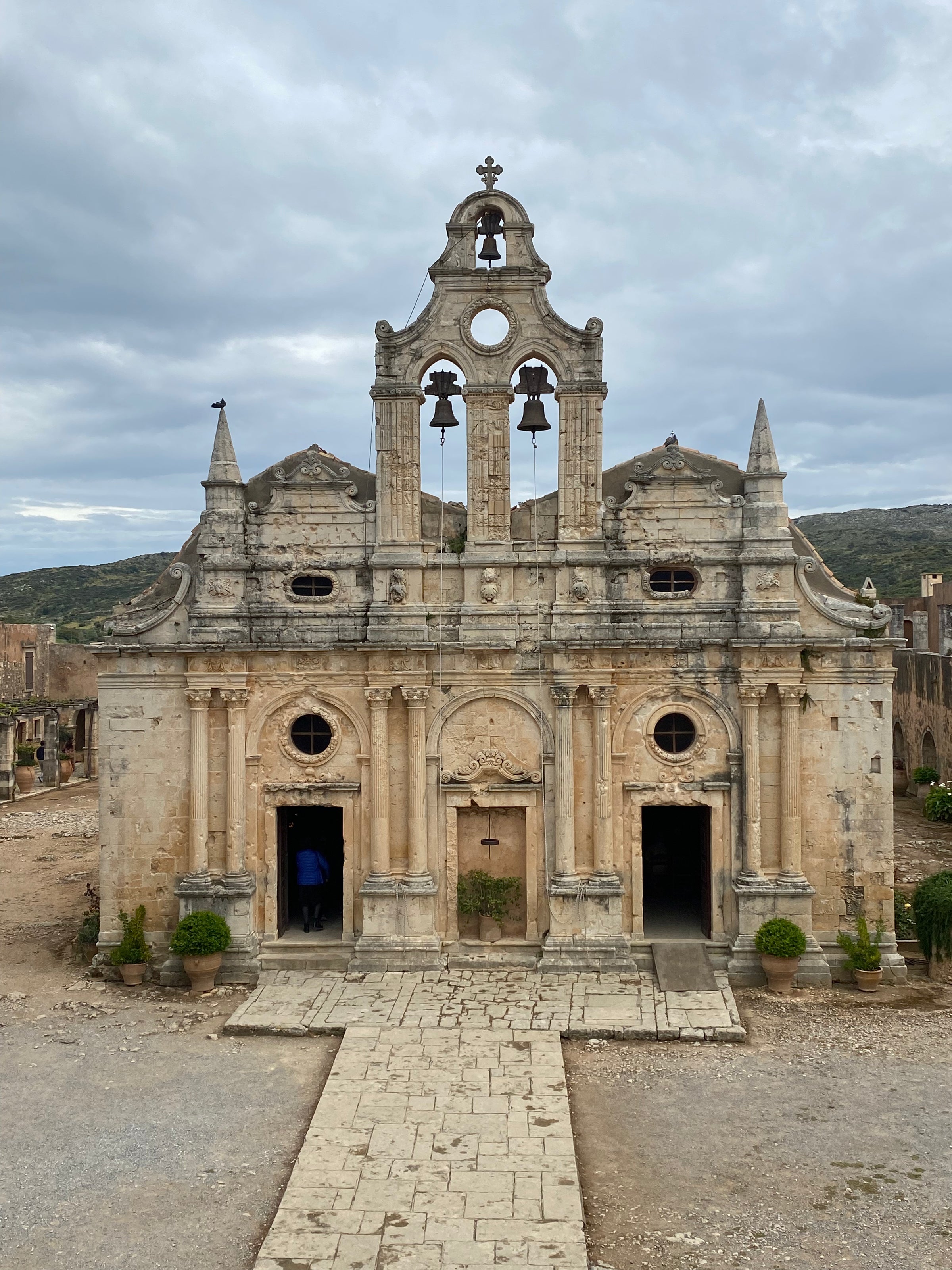 A historic stone church with two bell towers and three arched doorways stands under a cloudy sky, surrounded by a rustic landscape and ancient ruins.