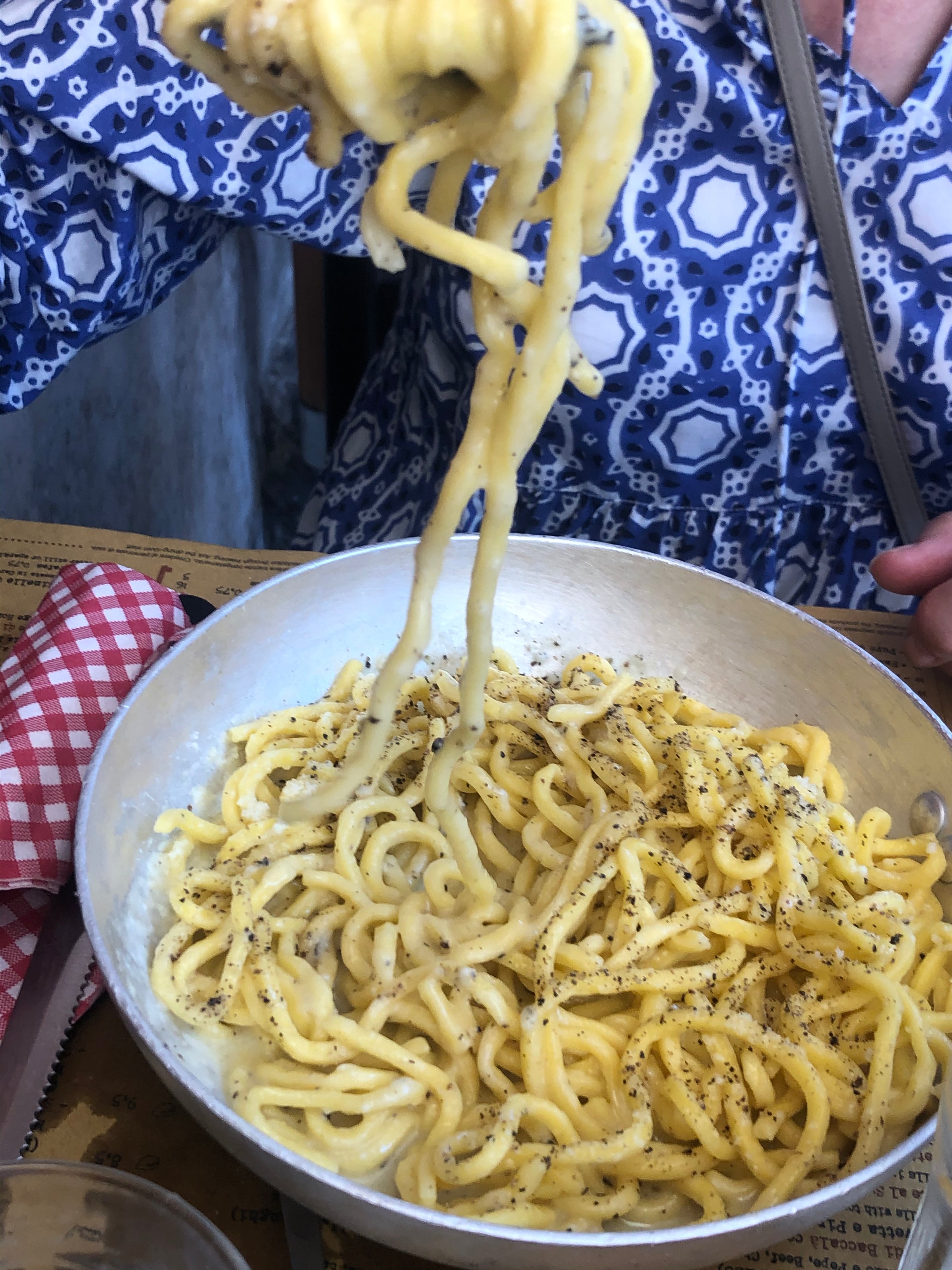 A person wearing a blue and white patterned shirt lifts a forkful of creamy pasta with black pepper from a metal bowl, with a red and white checkered napkin nearby.