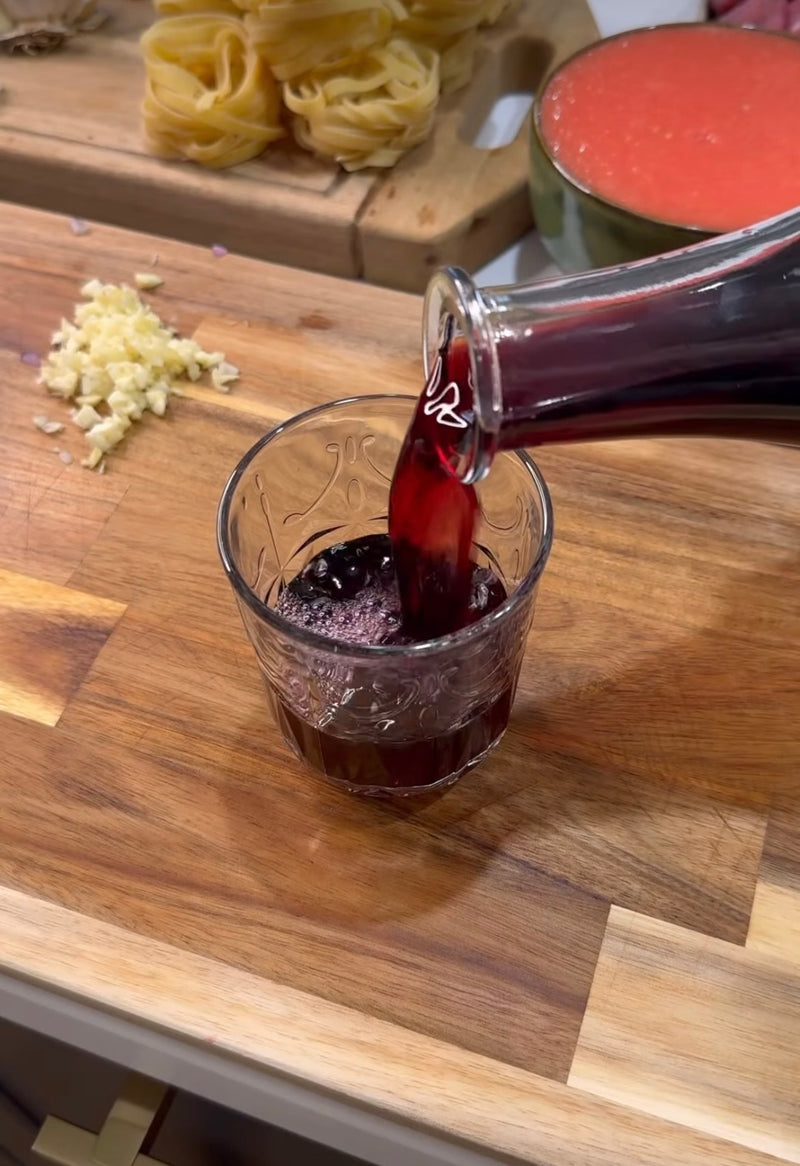 A hand pours red wine from a glass carafe into a decorative glass on a wooden counter. Chopped garlic, pasta nests, and a bowl of tomato sauce are visible in the background.