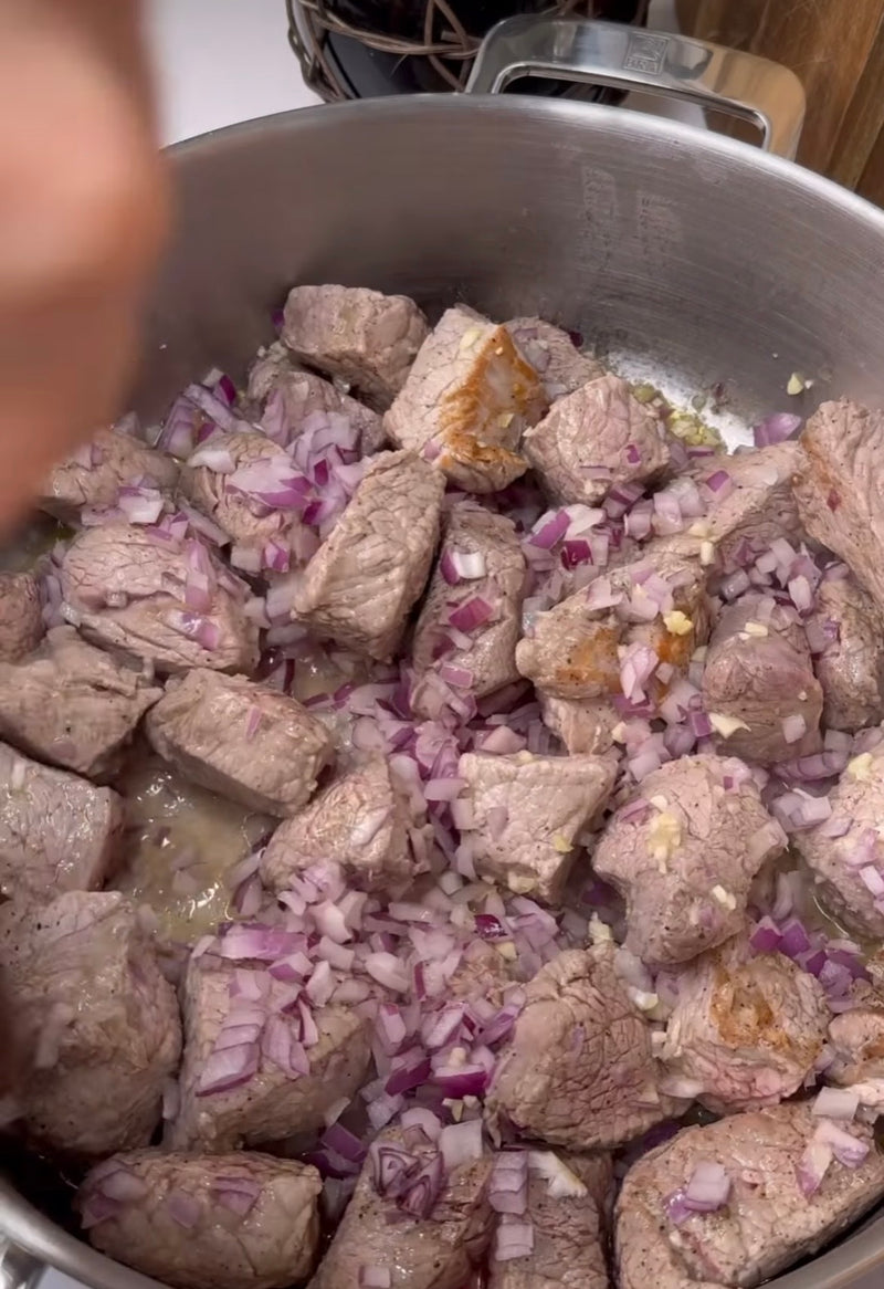 Chunks of beef are being browned in a pot with finely chopped red onions and minced garlic, starting to cook together.