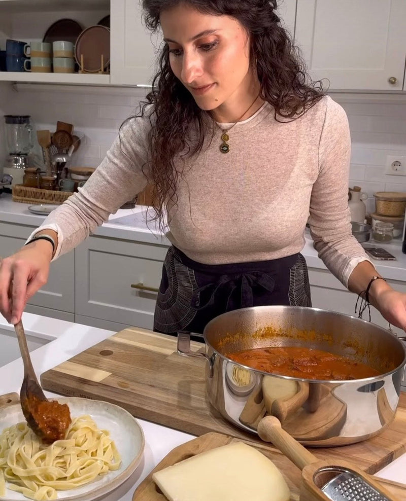 A woman in a kitchen spoons tomato sauce from a pot onto a plate of fettuccine pasta. Slices of cheese and a grater are on the counter nearby. Shelves and kitchenware are visible in the background.