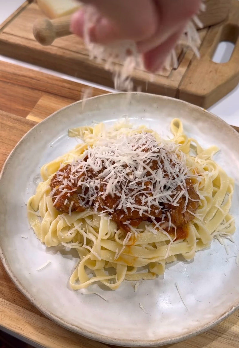 A close-up of a hand sprinkling grated cheese over a plate of tagliatelle pasta topped with tomato sauce, on a wooden surface with a cutting board in the background.