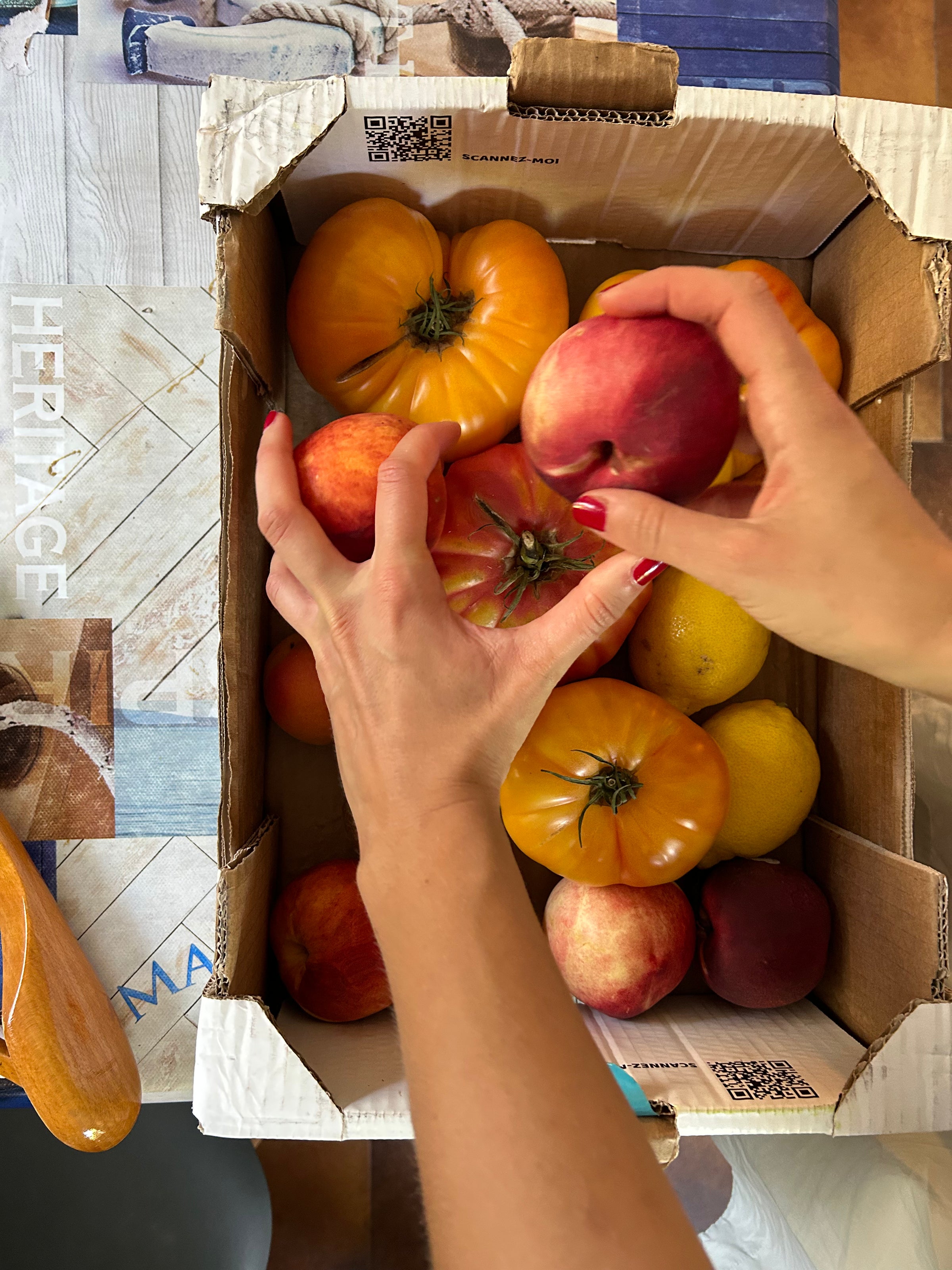 A person with red nail polish holds a peach and an heirloom tomato over a cardboard box filled with yellow tomatoes, peaches, and apples on a patterned table.