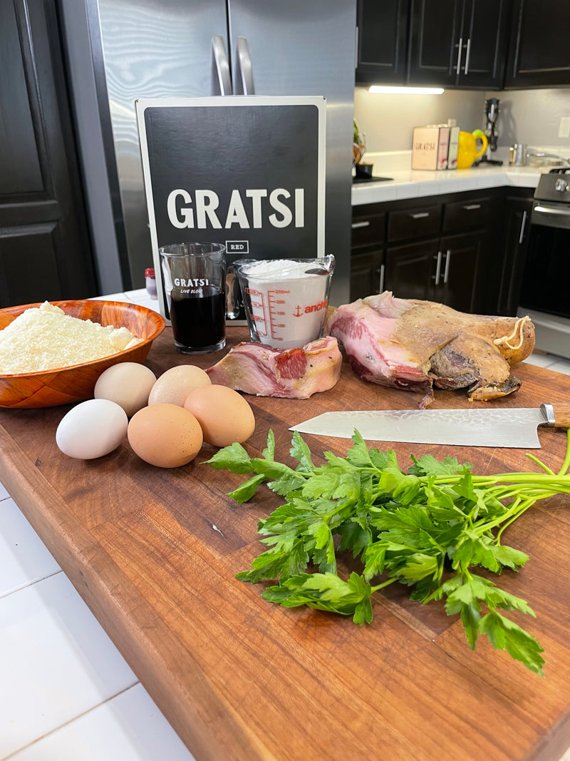 A wooden kitchen counter with eggs, parsley, pancetta, grated cheese, measuring cup, coffee cup, and a GRATS! sign in the background. A modern kitchen with dark cabinets is visible behind the counter.
