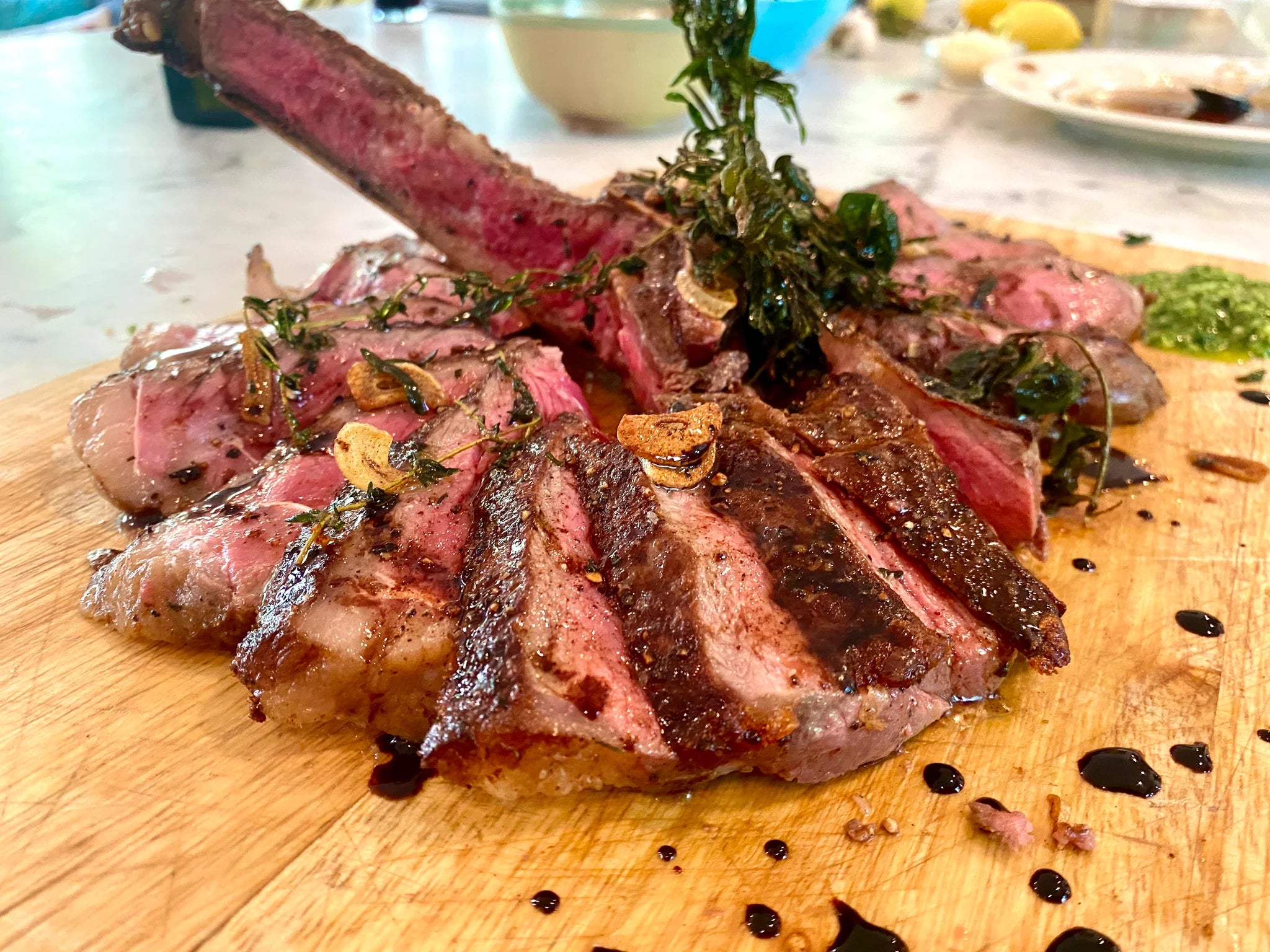 Sliced medium-rare tomahawk steak on a wooden cutting board, garnished with fried garlic, herbs, and a drizzle of sauce, with a blurred table setting in the background.