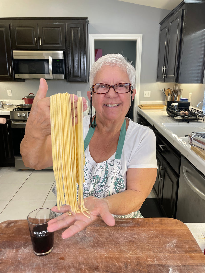 An older woman in glasses and an apron smiles while holding up freshly made pasta in a kitchen. A small glass of red wine sits on the wooden counter in front of her.