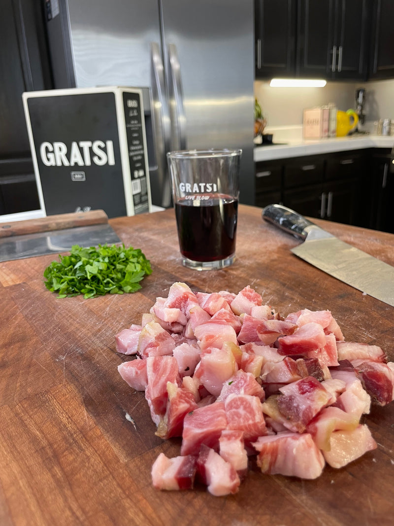 Diced pancetta on a wooden cutting board with chopped herbs, a knife, a glass of red wine, and a Gratsi wine box in a modern kitchen setting.
