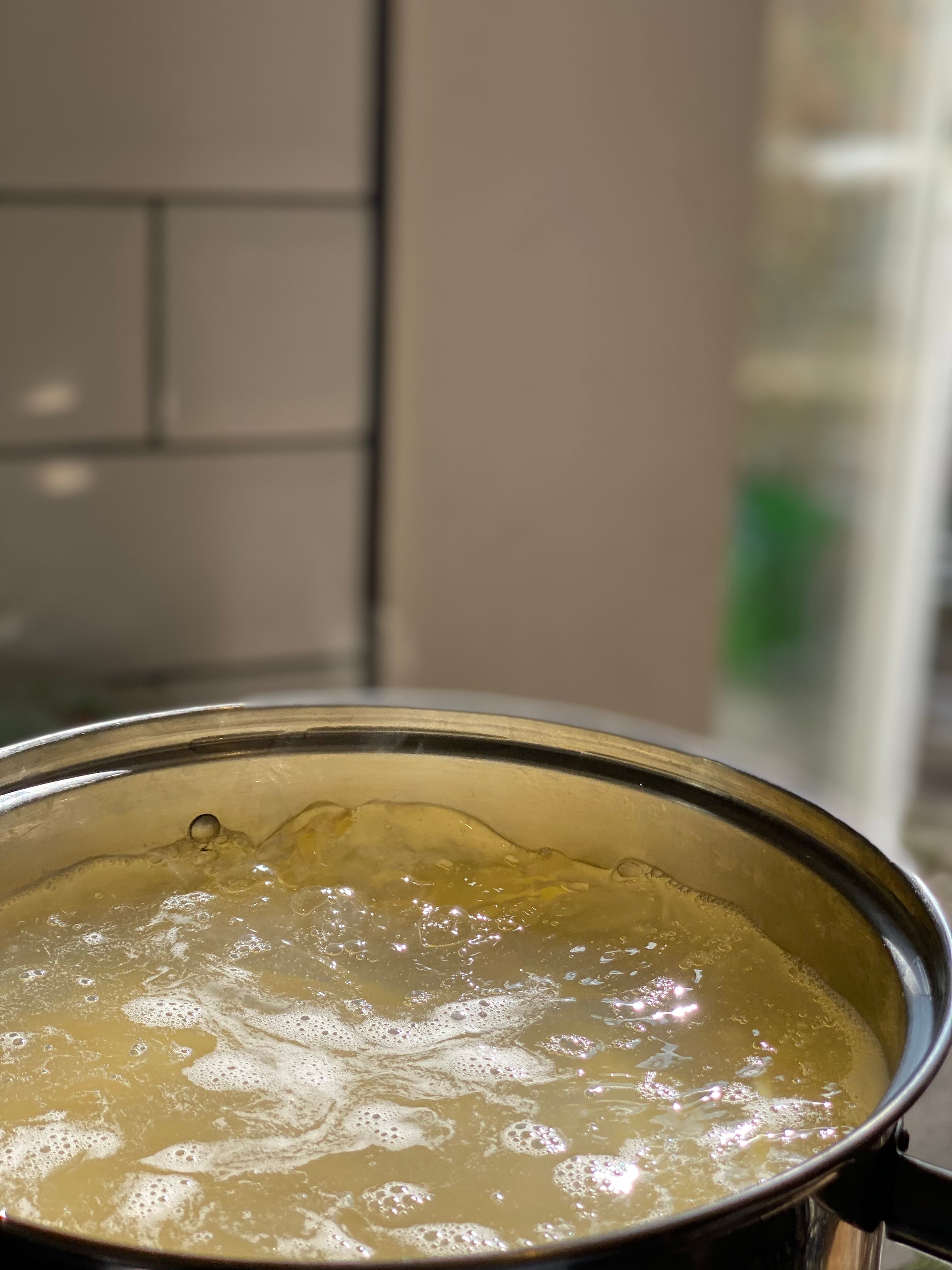 A close-up of a stainless steel pot filled with boiling water and partially submerged pasta, with steam rising. The background is out of focus, showing part of a tiled kitchen wall and window.
