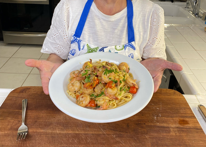 A person in a white eyelet shirt and blue apron holds a plate of shrimp pasta with cherry tomatoes and herbs over a wooden surface in a kitchen. A fork rests on the cutting board.
