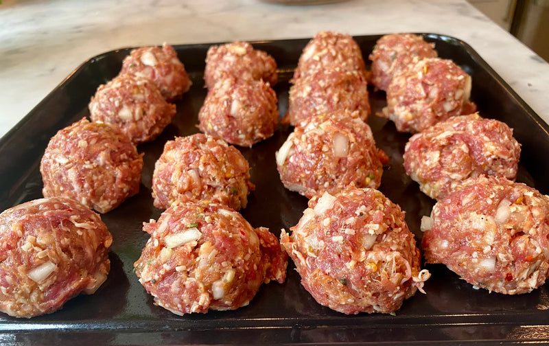 Raw meatballs mixed with chopped onions and seasonings are arranged on a black baking tray, ready to be cooked. The tray sits on a light-colored kitchen counter.