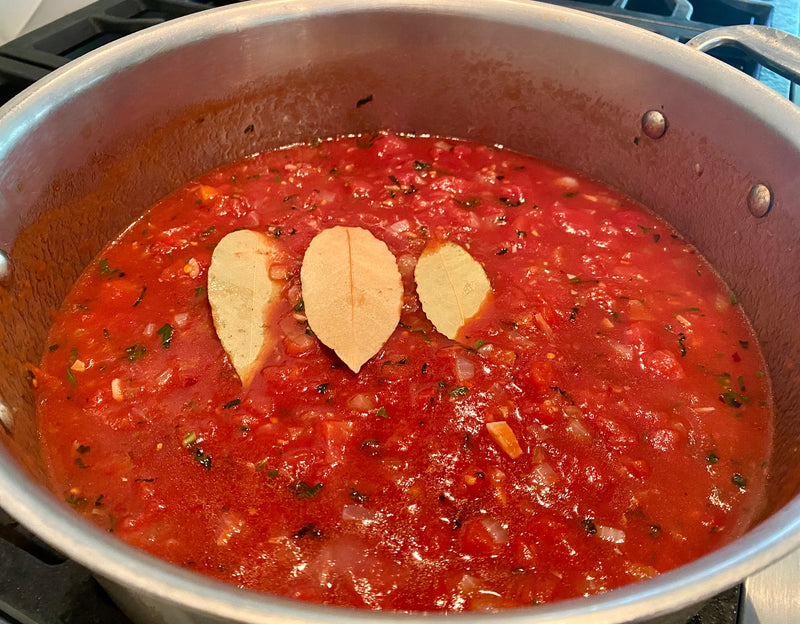 A pot of tomato sauce with visible herbs, onions, and diced vegetables is simmering on a stove. Three bay leaves are placed on top of the sauce.