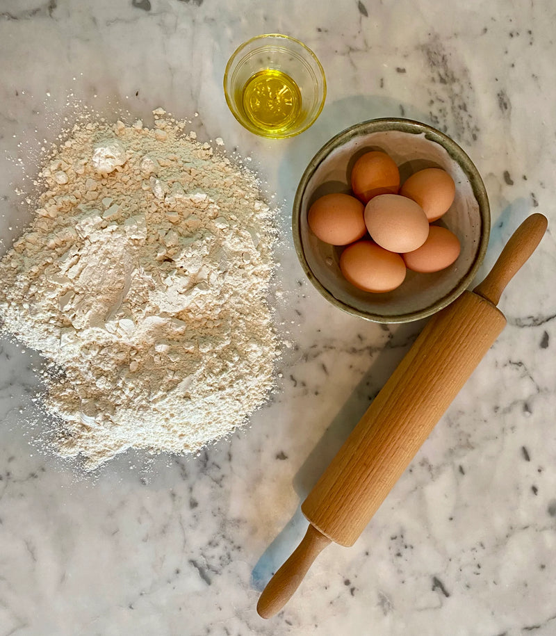 A pile of flour, a small bowl of olive oil, a bowl with six eggs, and a wooden rolling pin are arranged on a marble countertop.