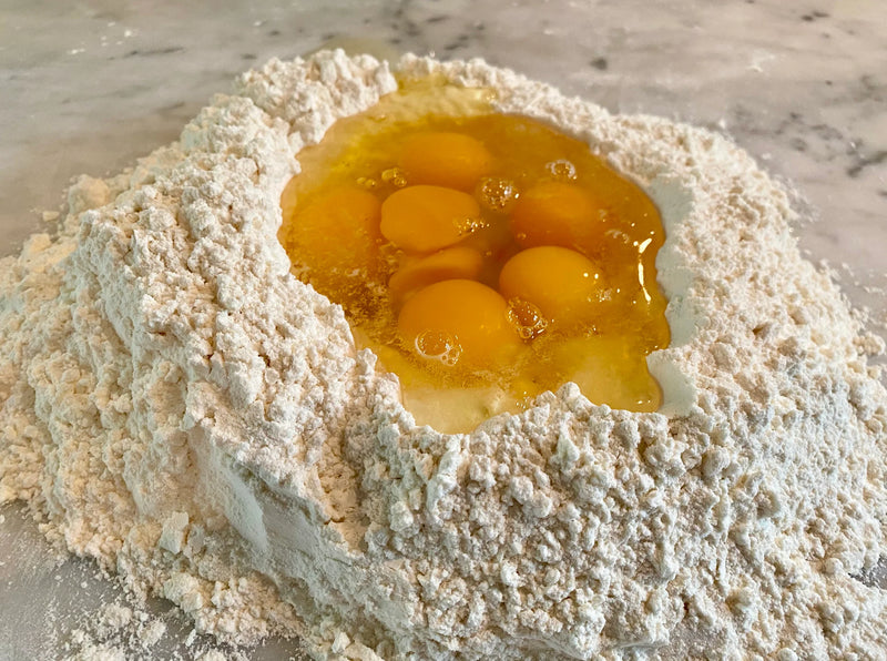 A mound of flour on a countertop with a well in the center holding several cracked eggs, ready for mixing, likely to make pasta dough.