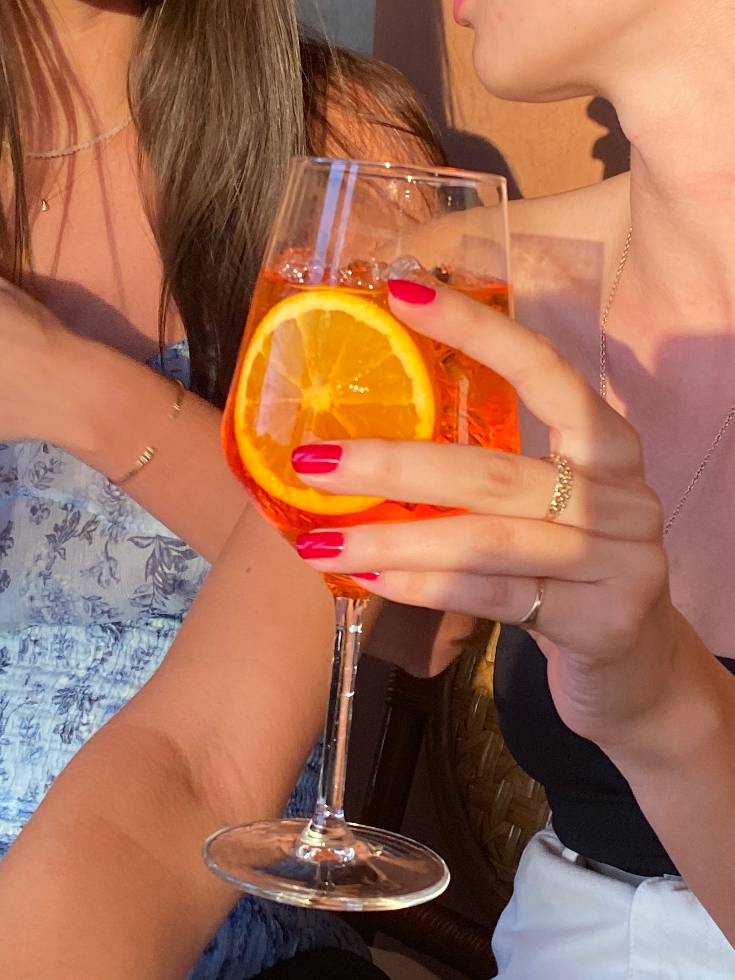Two women sit together; one holds a stemmed glass filled with an orange cocktail, garnished with an orange slice. She has pink nail polish and wears rings. Sunlight casts warm shadows on their skin.