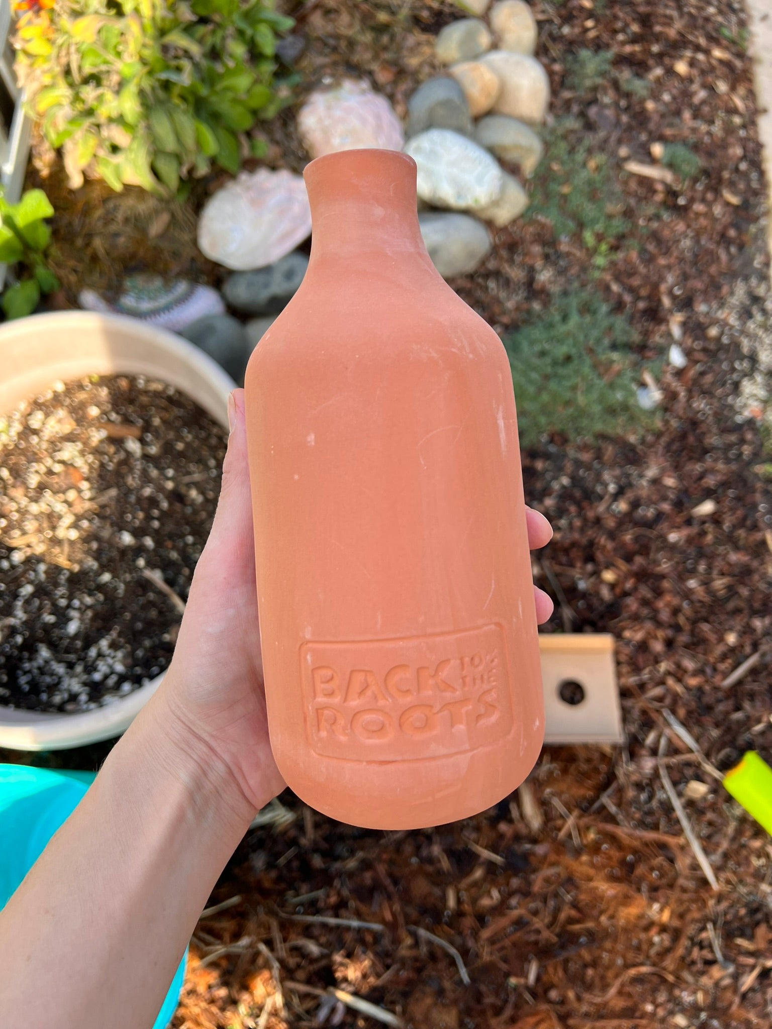 A hand holds a terracotta watering bottle stamped with Back to the Roots in a garden with soil, rocks, and plants visible in the background.