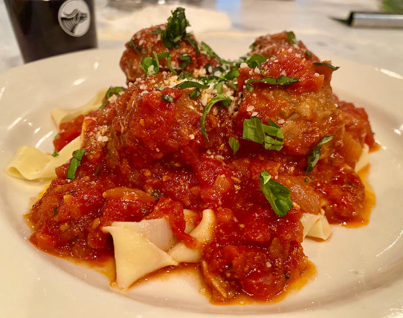 A plate of pappardelle pasta topped with meatballs, chunky tomato sauce, grated cheese, and fresh basil. The dish is served on a white plate with a dark cup in the background.