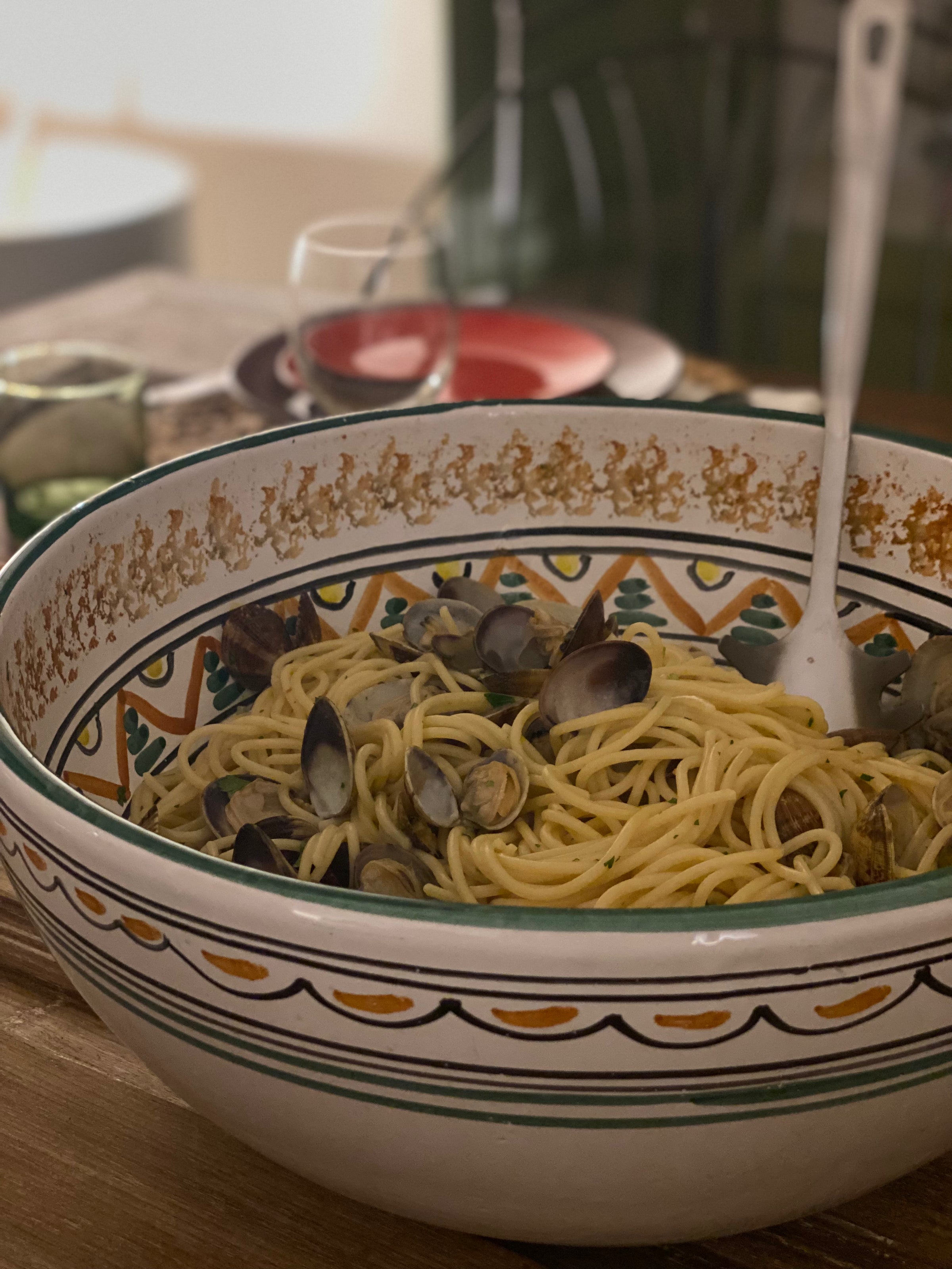 A large decorative bowl filled with spaghetti and clams sits on a table, with a serving spoon inside. In the background, there are plates, a glass, and a candle.