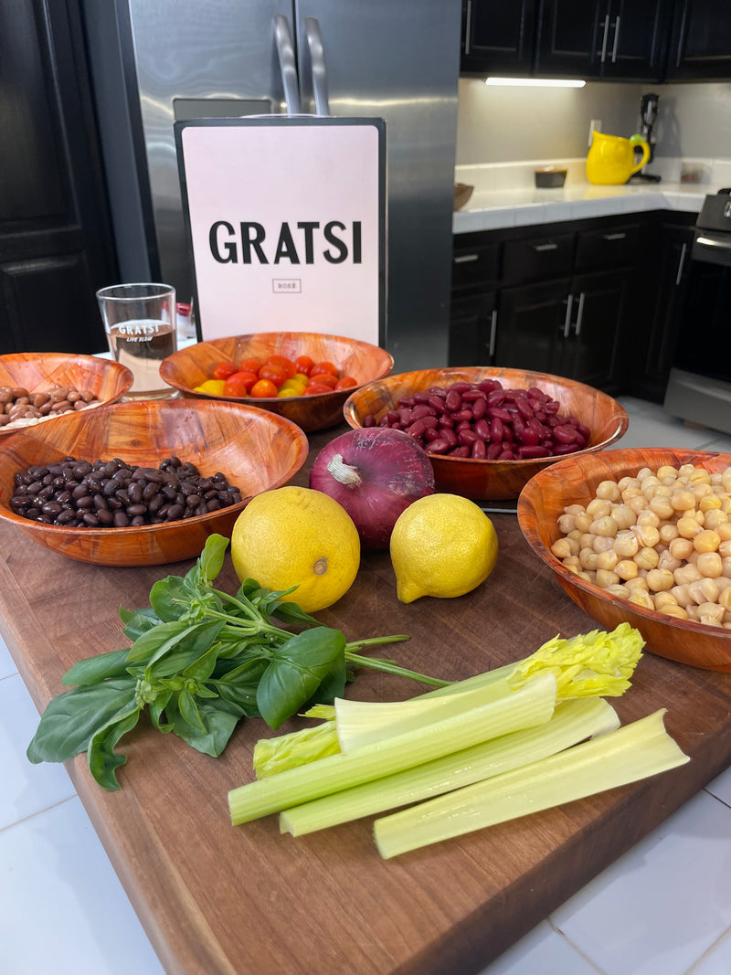 A wooden cutting board with celery, lemons, red onion, basil, and bowls of chickpeas, black beans, kidney beans, cherry tomatoes, and olives, set in a modern kitchen with a GRATSI sign in the background.
