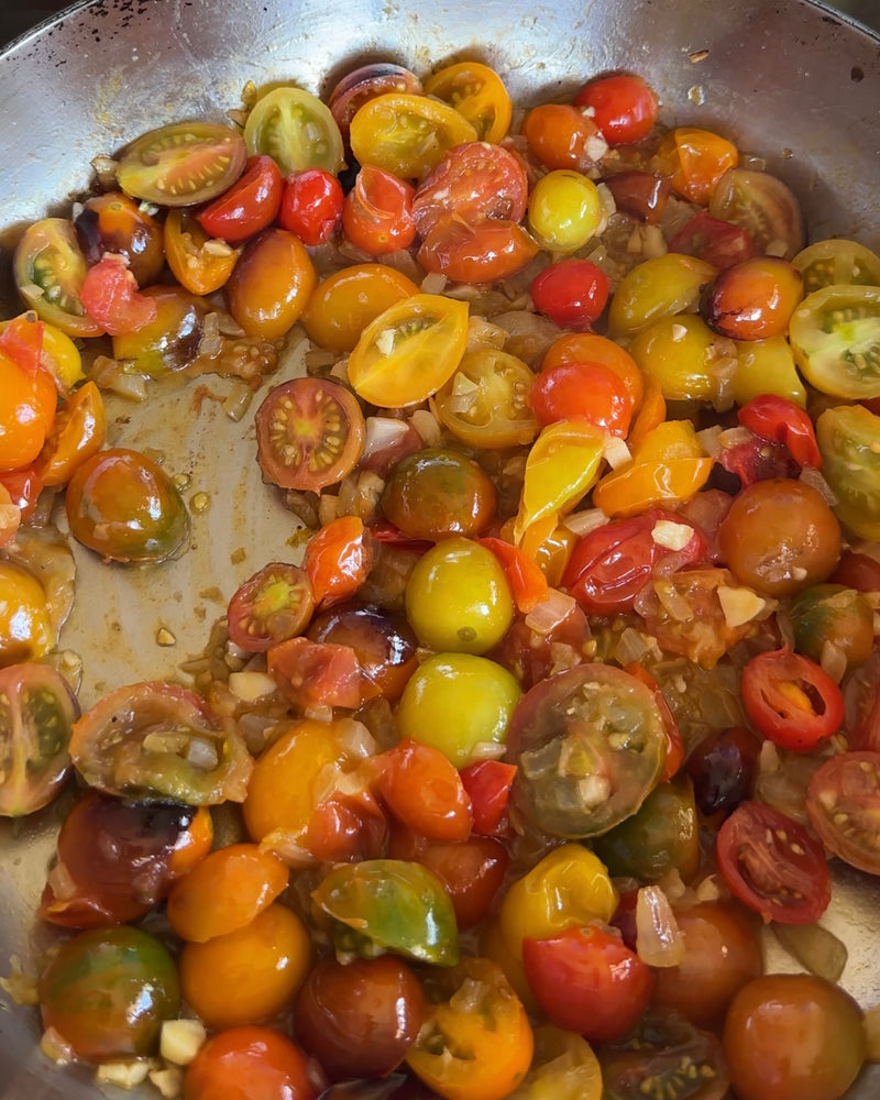 A pan filled with chopped, colorful cherry tomatoes and diced onions being sautéed. The tomatoes are red, yellow, orange, and green, creating a vibrant mixture.
