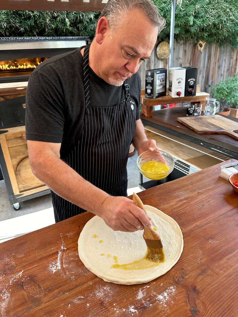 A man in a black apron brushes olive oil onto a round pizza dough on a wooden counter, with a wood-fired oven and pizza ingredients visible in the background.