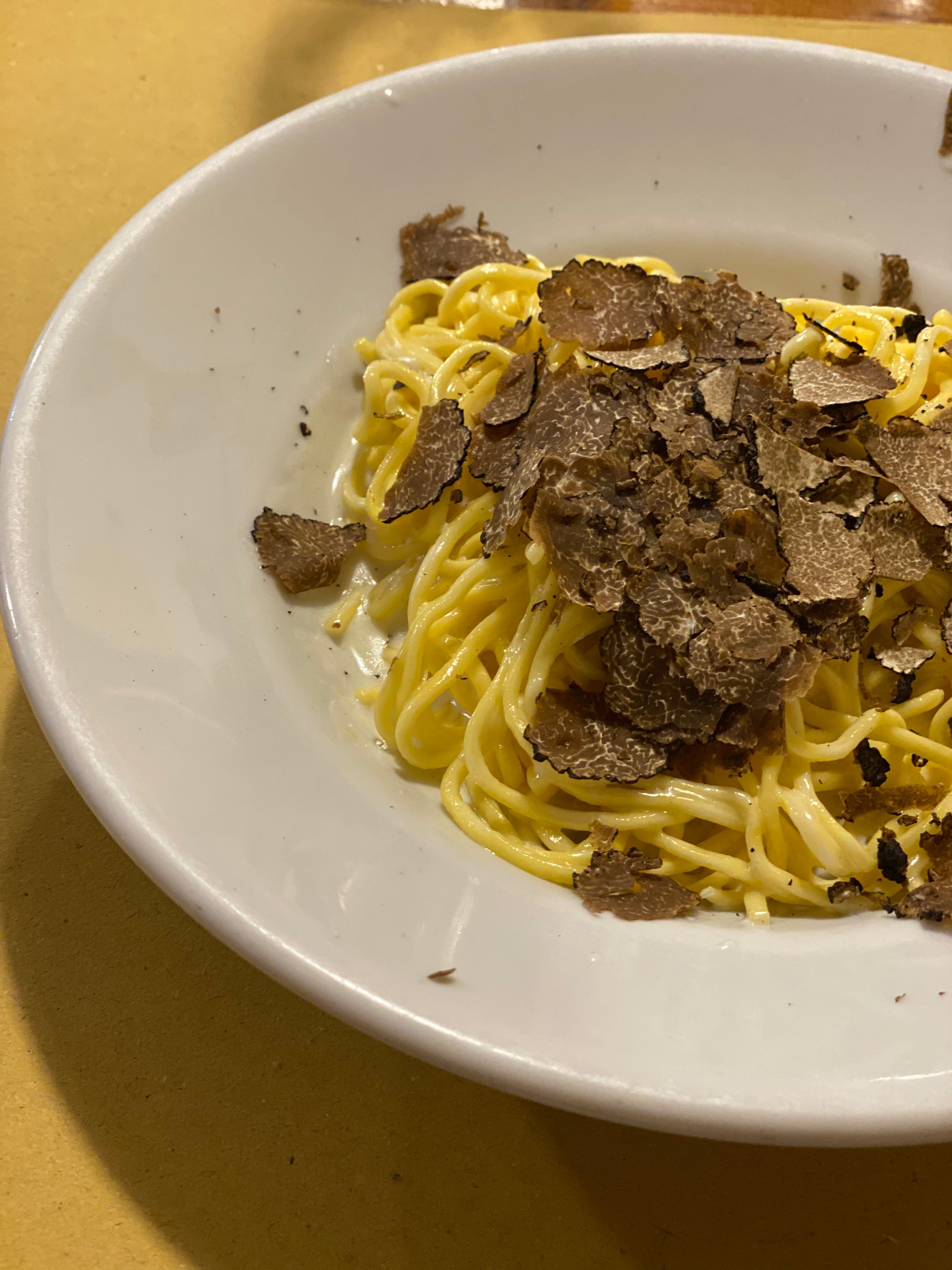 A white plate filled with creamy pasta topped with generous shavings of black truffle, placed on a light brown table.