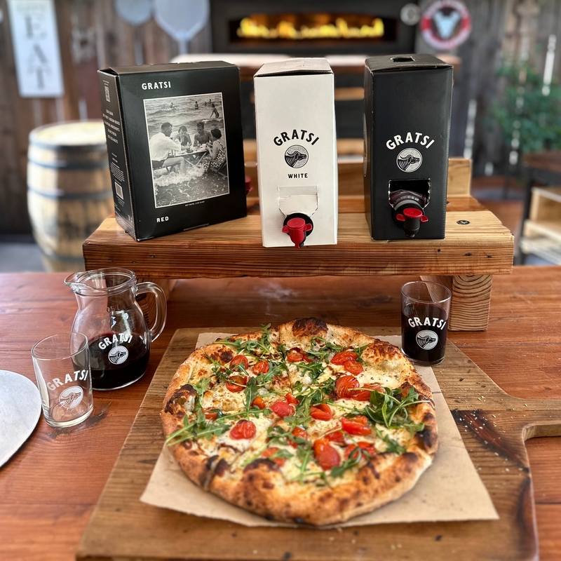 A rustic table with a fresh pizza topped with arugula and cherry tomatoes, a glass pitcher and two glasses of red wine, and three boxes of Gratsi wine (red, white, and black) in the background.