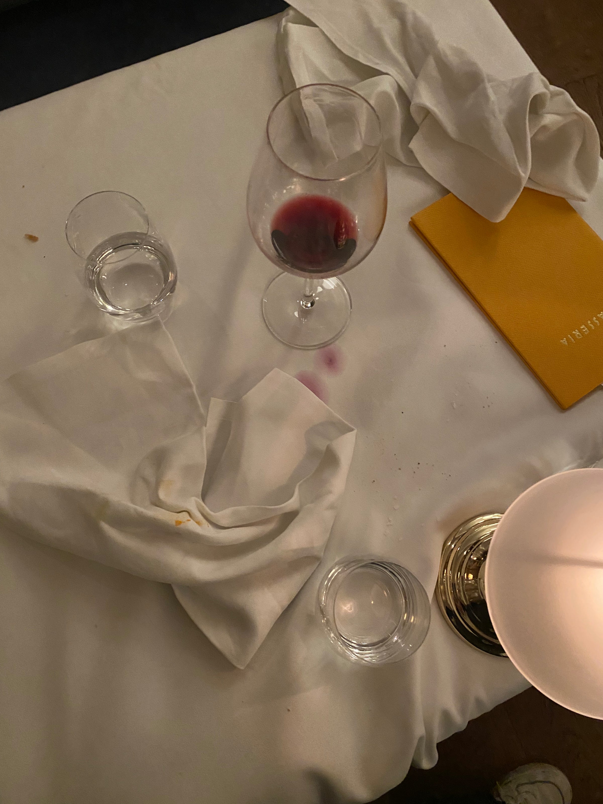 A dimly lit table with two glasses of water, a glass of red wine with a wine stain on the white tablecloth, a yellow menu, crumpled napkins, and a lamp in the corner.