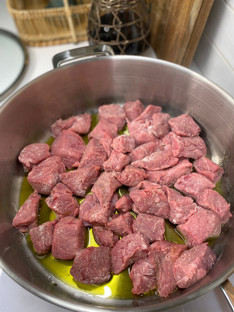 Chunks of raw beef are spread in a layer in a stainless steel pot with a small pool of olive oil, ready for browning. The background shows a woven basket and kitchen items.