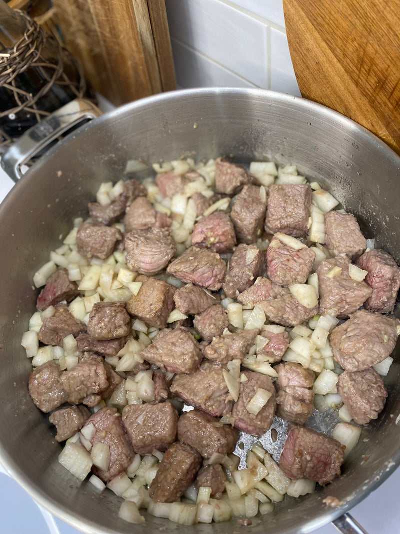 Chunks of beef and chopped onions are cooking in a large stainless steel pot on a stovetop, surrounded by kitchen utensils and a wooden cutting board.
