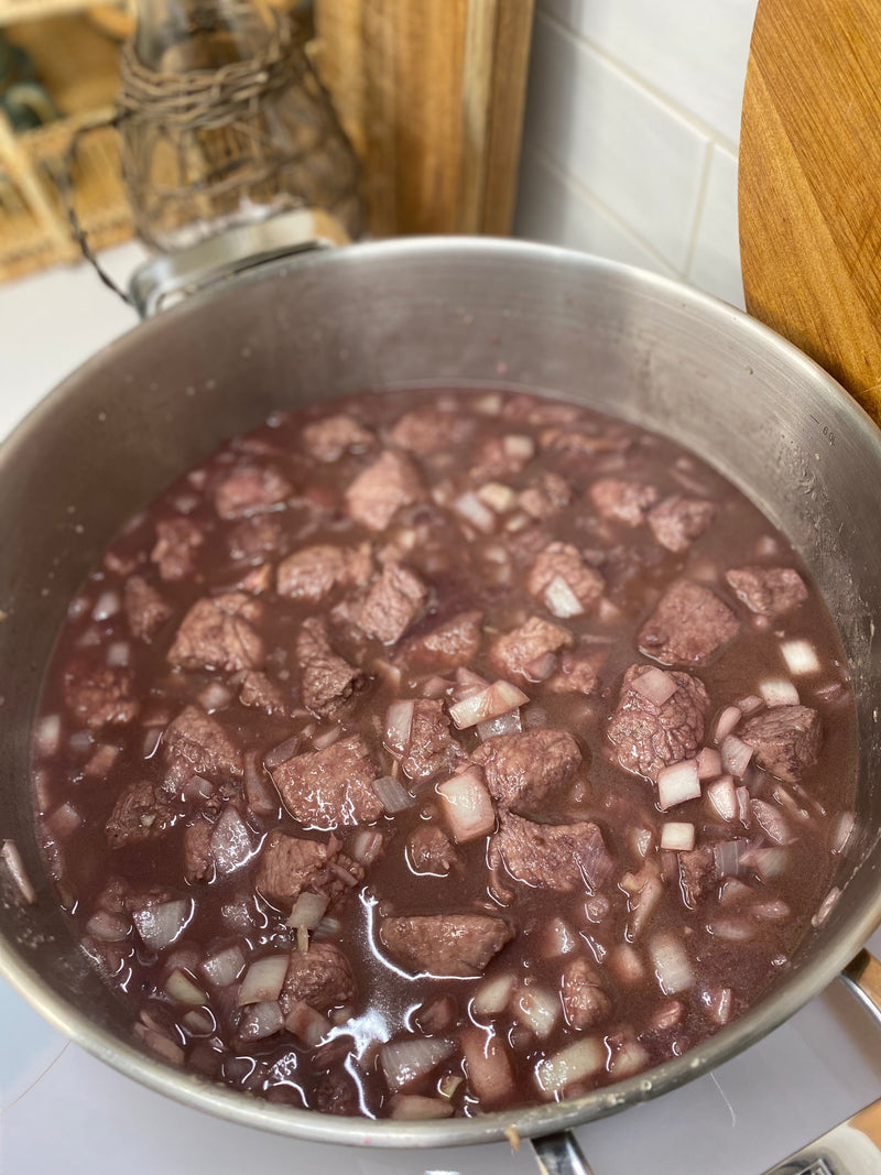 A large pot filled with chunks of beef and diced onions simmering in a dark liquid, likely a stew or braise, sits on a kitchen stove.