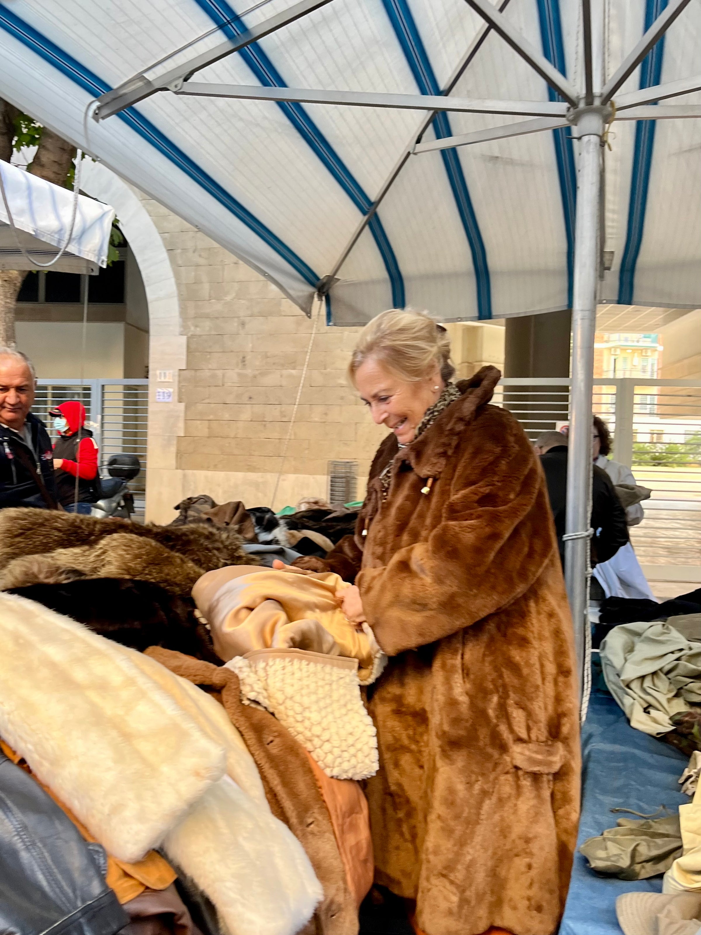 A woman in a large brown fur coat smiles while looking through a pile of coats at an outdoor market stall under a striped canopy. Other people and buildings are visible in the background.