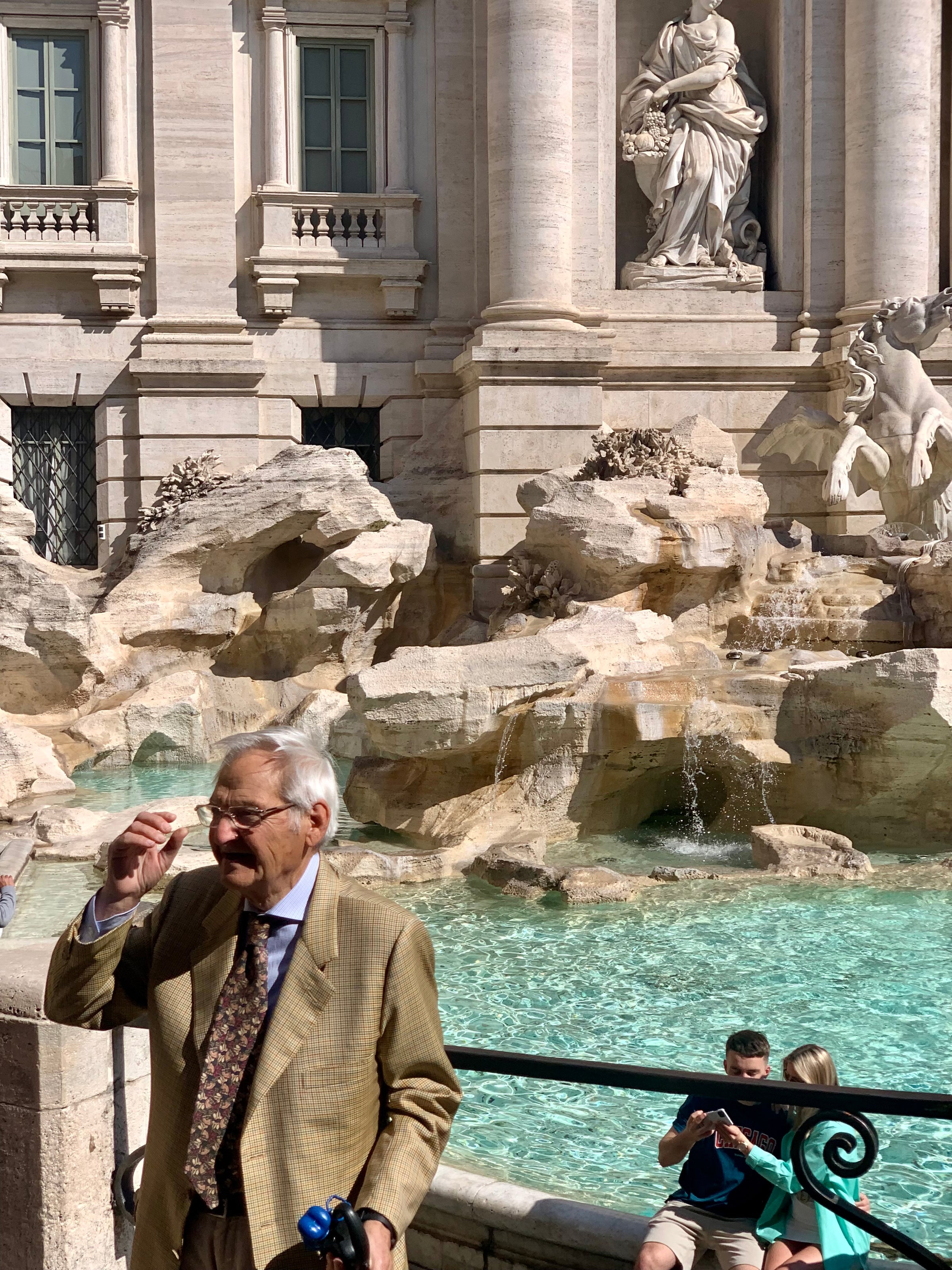An older man in a tan checkered suit holds his glasses near the Trevi Fountain in Rome. Behind him, tourists sit by the turquoise water and ornate statues and stonework decorate the fountain.