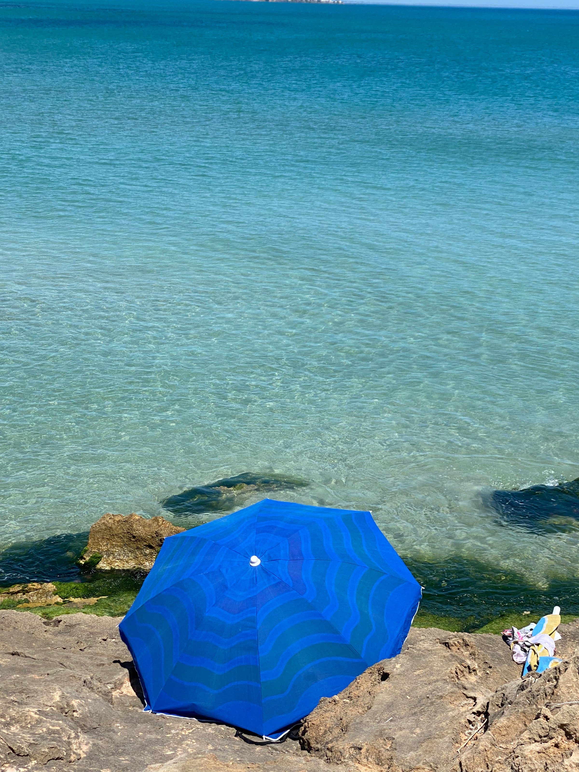 A bright blue umbrella sits on rocky shore beside clear, turquoise water. A small colorful towel and flip-flops are nearby on the rocks. The calm sea stretches towards the horizon under a clear sky.