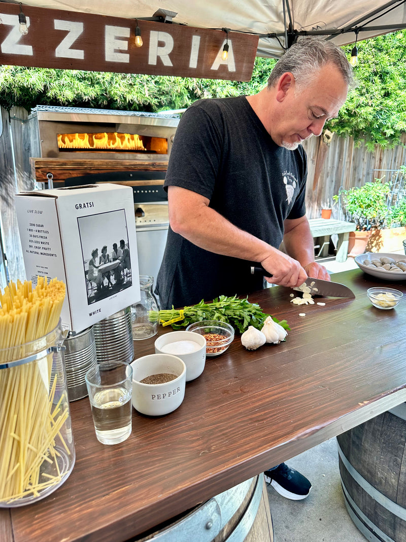 A man slices garlic on a wooden counter outdoors, surrounded by fresh herbs, a jar of spaghetti, seasonings, and a glass of white wine. A wood-fired oven and a Pizzeria sign are in the background.
