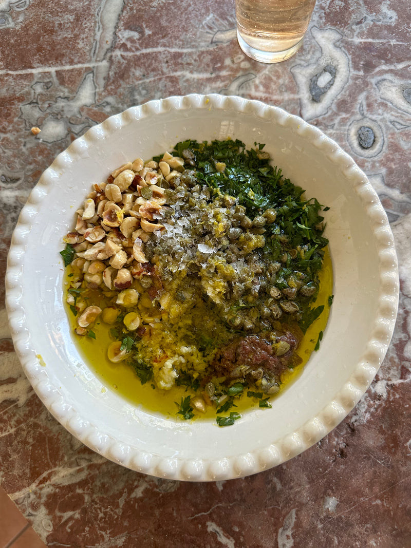 A white bowl on a marbled surface holds chopped parsley, capers, toasted hazelnuts, grated lemon zest, anchovies, olive oil, and cracked pepper, ready to be mixed. A glass of light-colored liquid is in the background.