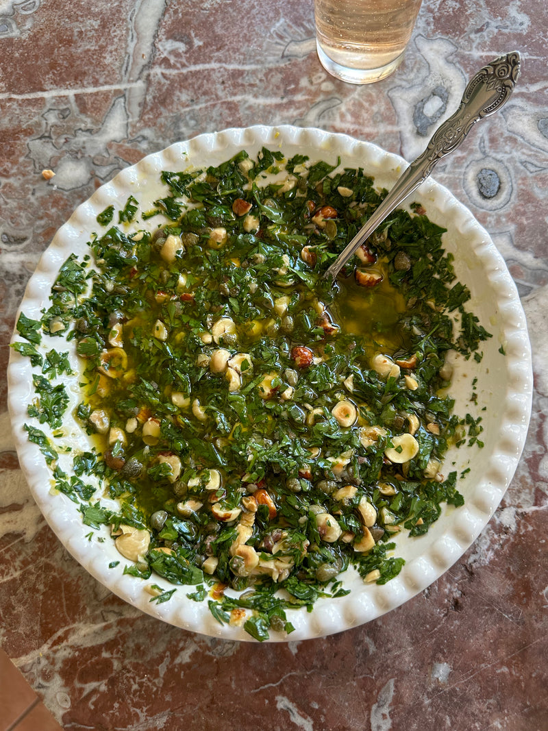A white bowl with a scalloped edge holds a green herb sauce with chopped nuts and oil; a spoon rests in the bowl, which sits on a marbled tabletop beside a glass of light-colored liquid.