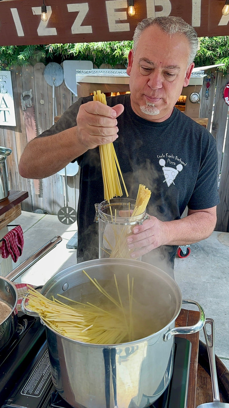 A man is holding uncooked spaghetti above a steaming pot, preparing to add it to boiling water. He is standing outdoors, wearing a black t-shirt with a small logo. Cooking utensils and kitchen items are visible in the background.