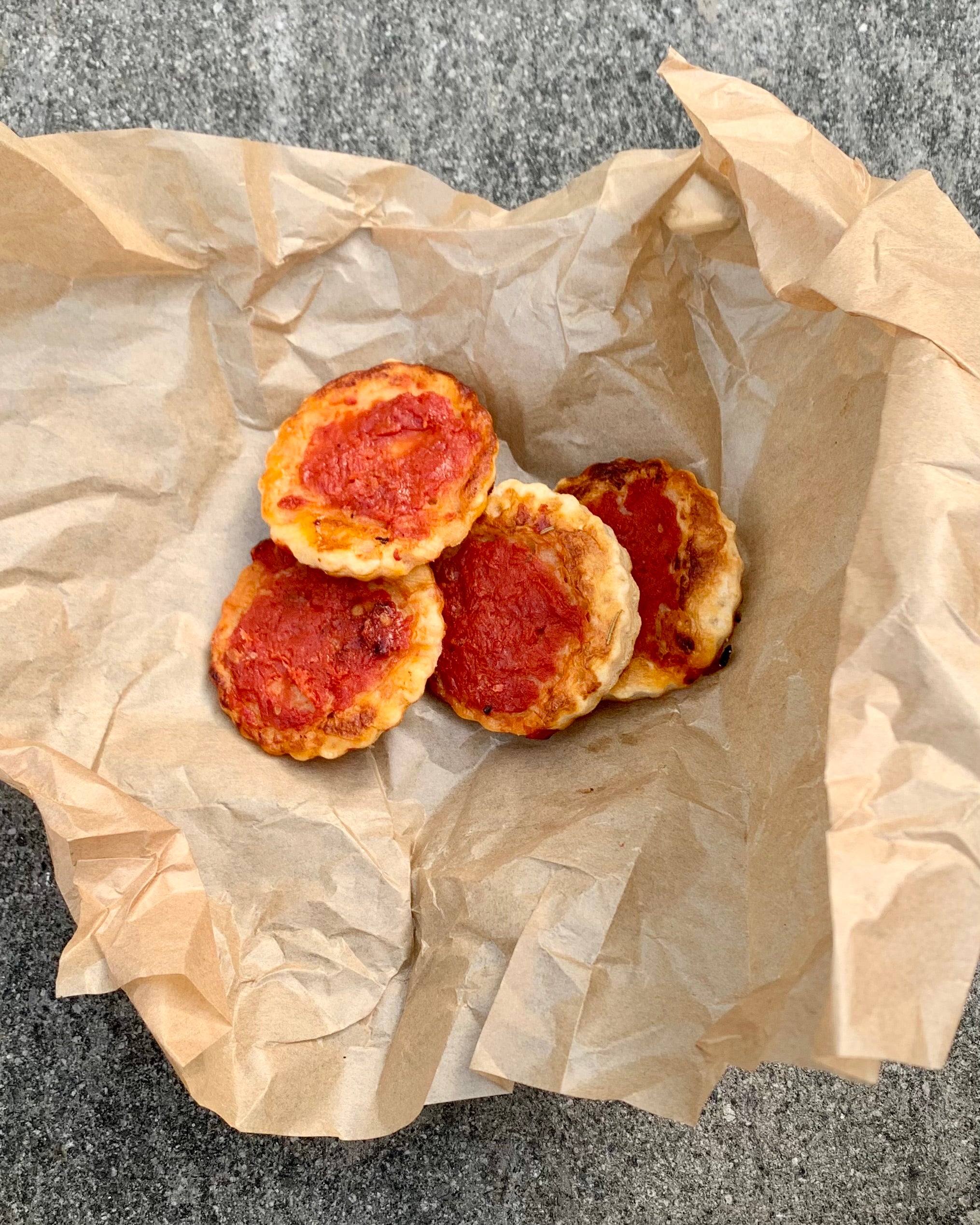 Four small pizza bites with tomato sauce and cheese are placed on crumpled brown parchment paper, resting on a gray surface.