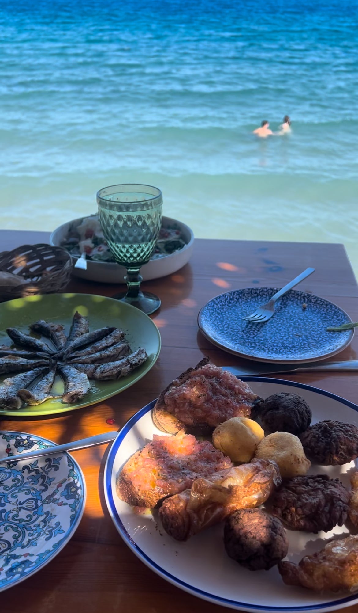 A seaside table set with plates of grilled sardines, roasted potatoes, bread, and a glass of green wine, with two people swimming in the blue ocean in the background.