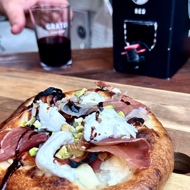 A close-up of a flatbread topped with prosciutto, cheese, pistachios, and balsamic glaze on a wooden board, with a hand holding a glass of red wine in the background next to a boxed wine dispenser.