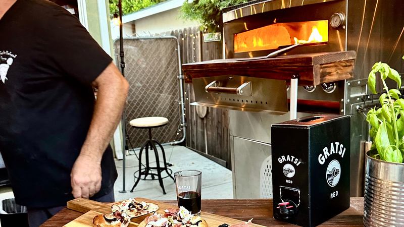 A person stands near an outdoor pizza oven with a fire inside. A wooden table holds bruschetta, a glass of red wine, a basil plant, and a Gratis Red wine box. String lights and a stool are in the background.