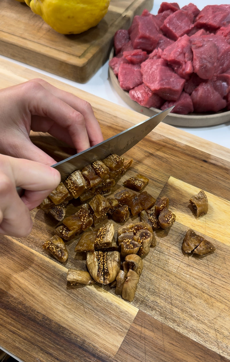 A person slicing dried figs on a wooden cutting board with a knife; chunks of raw red meat and a yellow fruit are visible in the background.