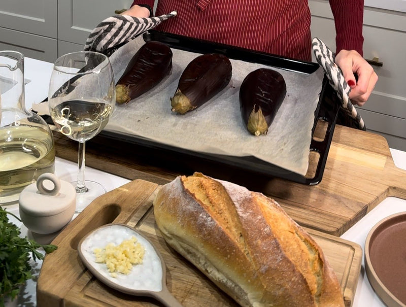 A person in a red apron holds a baking tray with three roasted eggplants on parchment paper. In the foreground are a loaf of bread, a plate with chopped garlic and sauce, a glass of white wine, and fresh herbs.