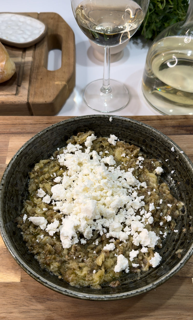 A bowl of roasted eggplant topped with crumbled feta cheese sits on a wooden table next to a glass of white wine, a wine carafe, and a cutting board with bread.