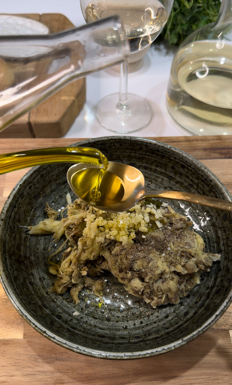A spoon pours golden olive oil over a bowl of shredded mushrooms and chopped garlic on a wooden board. In the background, a wine glass and a carafe of white wine are visible.