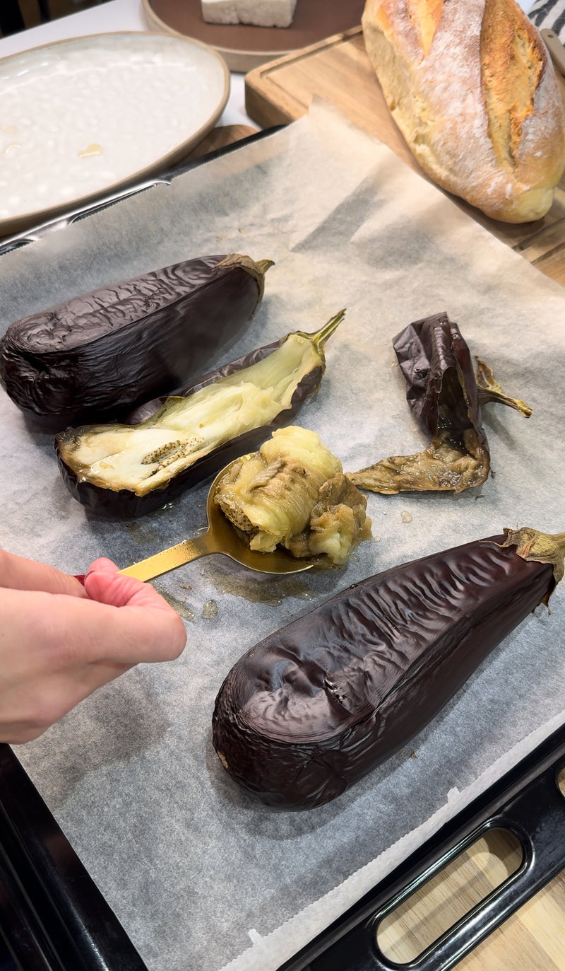 A hand uses a spoon to scoop out the roasted flesh from a baked eggplant on a parchment-lined baking tray, with two other roasted eggplants and a piece of bread nearby.