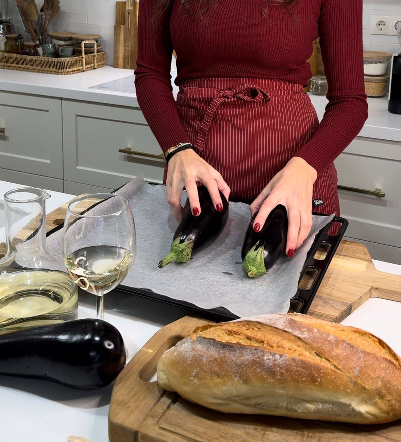 A woman in a red top and apron places eggplants on a baking tray lined with parchment paper. Nearby are a loaf of bread, a glass of white wine, and kitchen utensils on a wooden countertop.