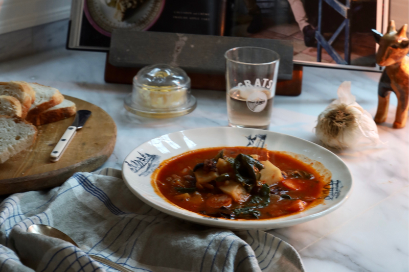 A bowl of vegetable soup with pasta sits on a table next to sliced bread, butter, a knife, a glass of water, and a decorative figurine, with a striped napkin in the foreground and books in the background.