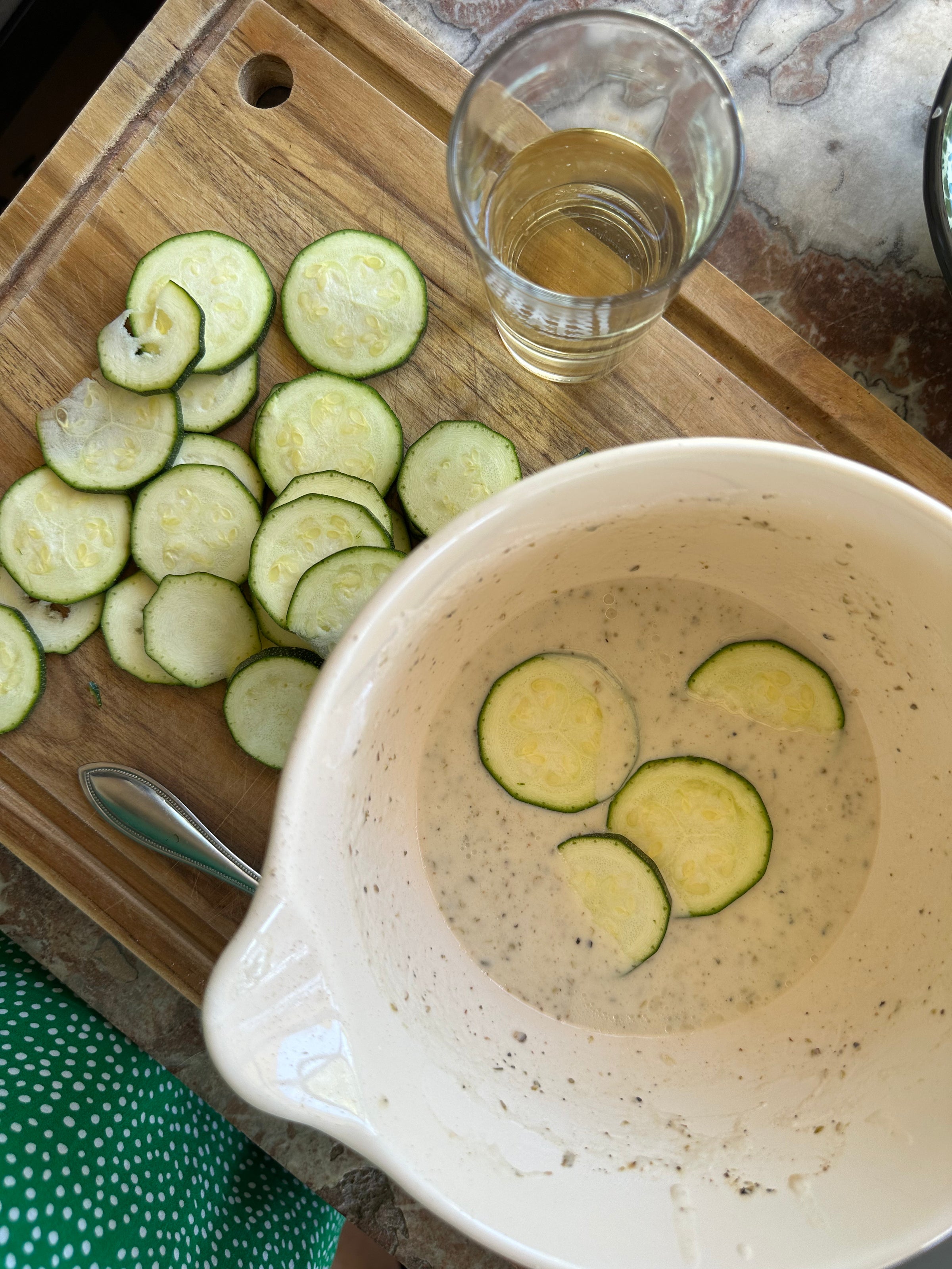 Sliced zucchini on a wooden cutting board next to a glass of liquid. A mixing bowl in the foreground contains zucchini slices soaking in a white batter. A spoon and part of a green polka-dot dress are visible.