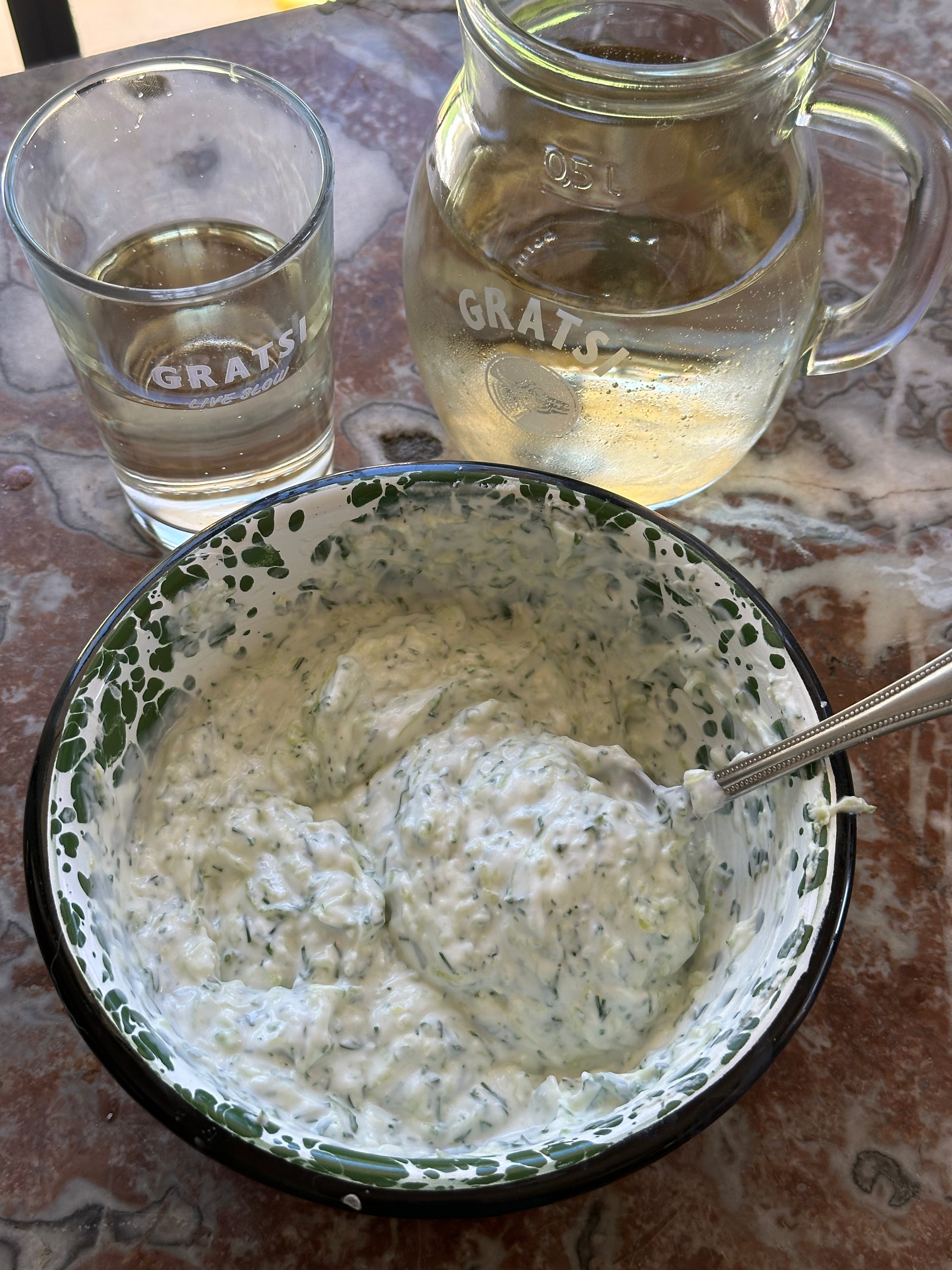 A bowl of creamy, herb-filled tzatziki with a spoon sits on a marble table next to a glass and a pitcher of chilled white wine, both labeled GRATSI.
